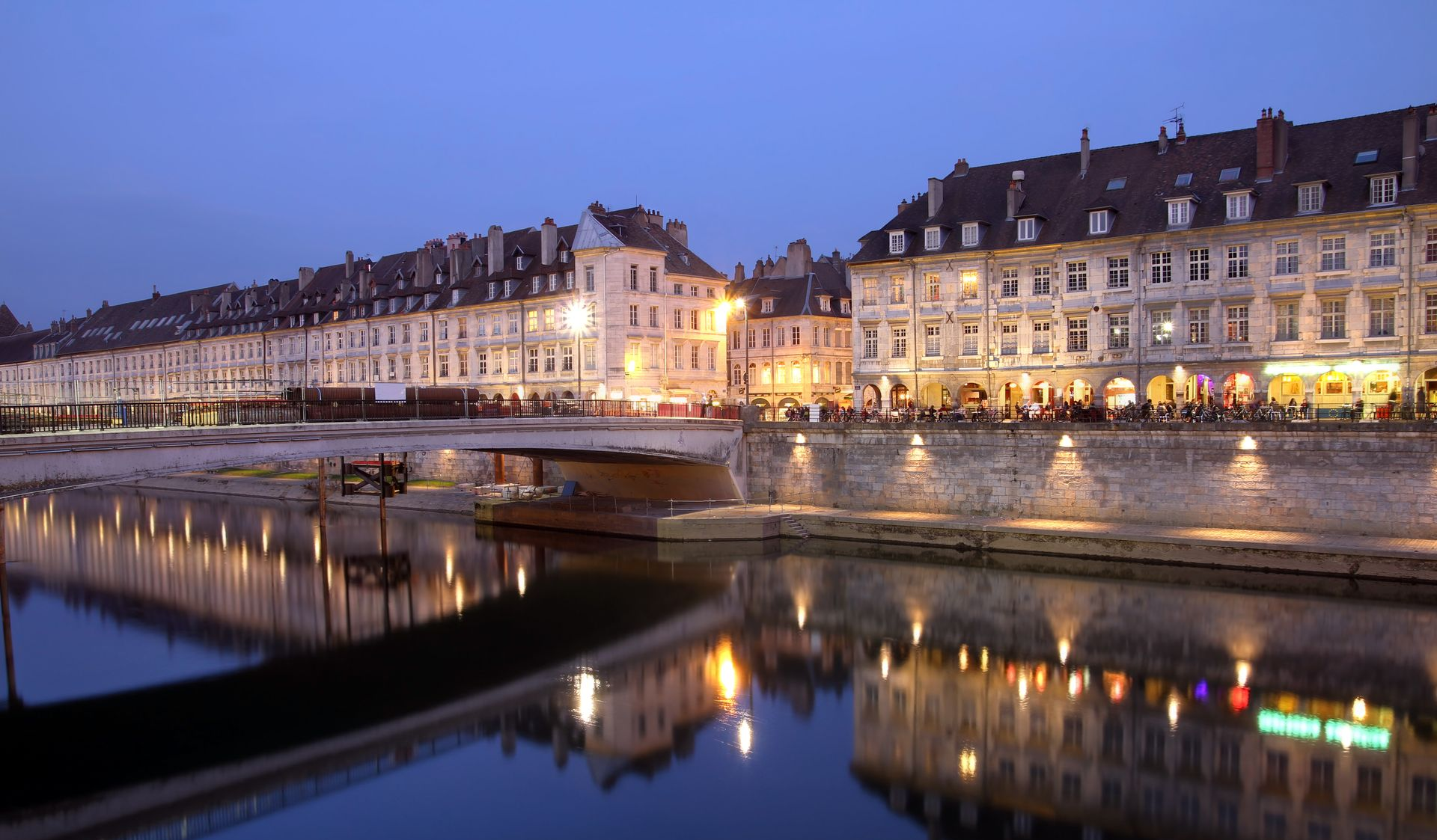 Vue nocturne de bâtiments historiques aux lumières chaudes se reflétant dans une rivière calme, devant un quai de pierre.