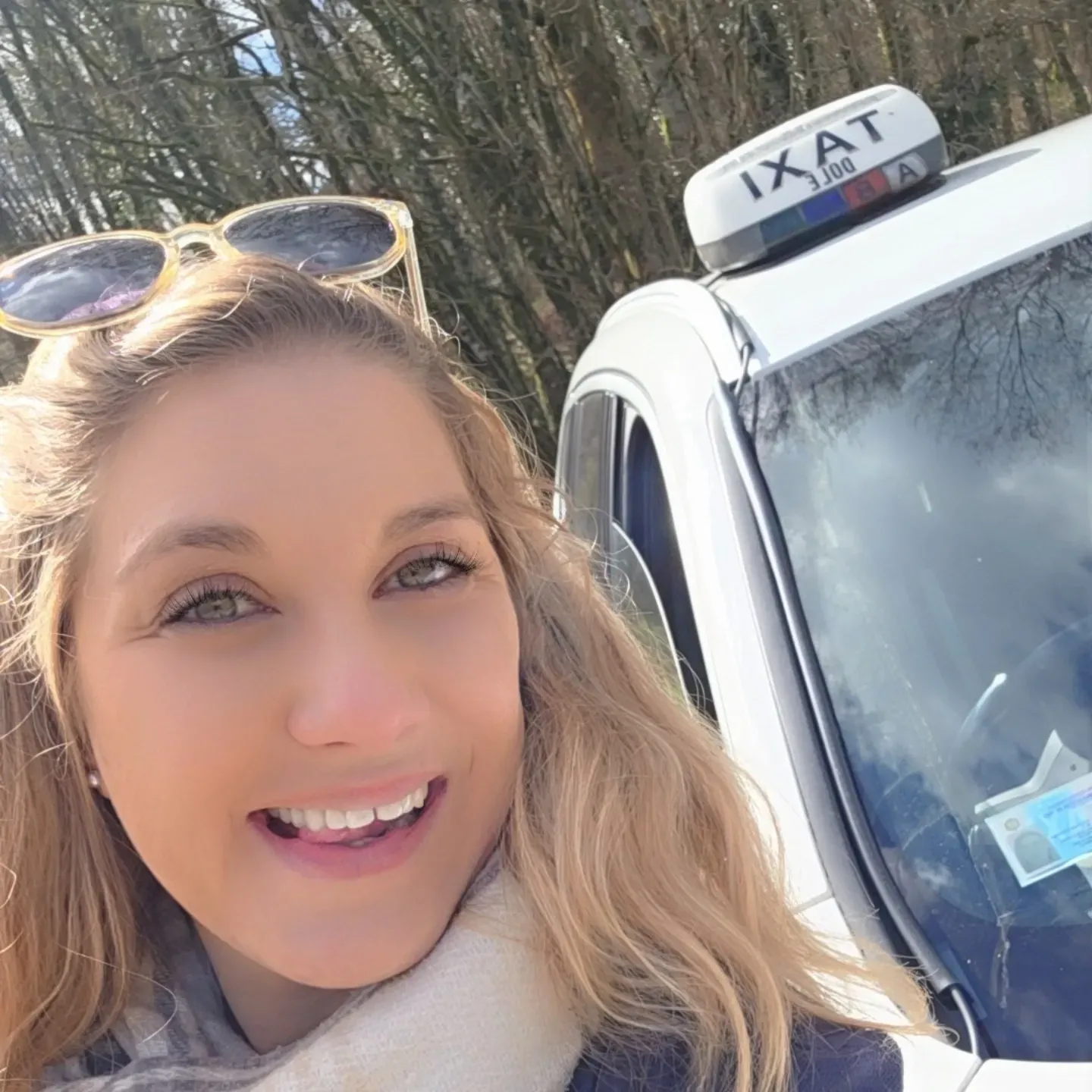 Une femme souriante, aux longs cheveux et portant des lunettes de soleil sur la tête, se tient devant un taxi.