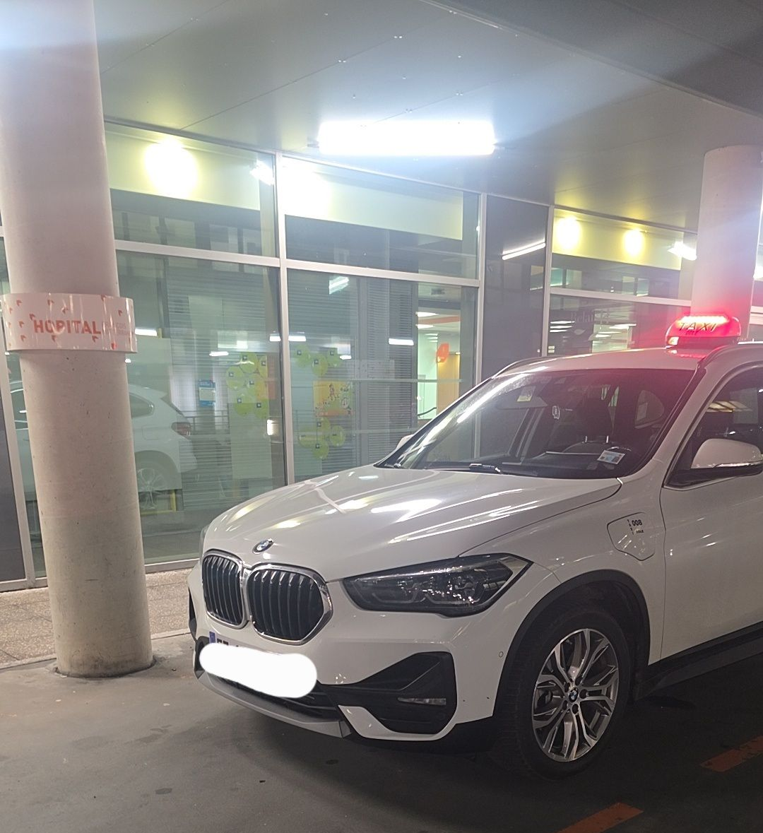 Un SUV BMW est garé de nuit près d'un pilier avec un gyrophare sur le toit, devant un hôpital aux portes vitrées.
