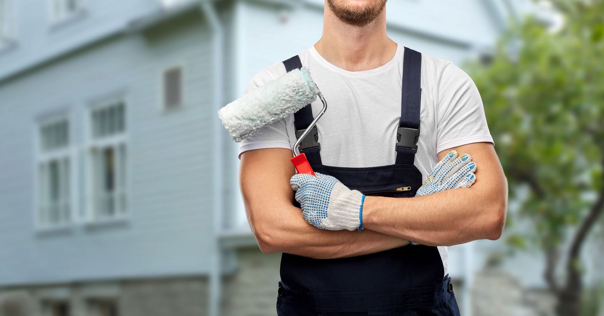 Ein Maler in Overalls hält eine Farbrolle und steht vor einem Haus.