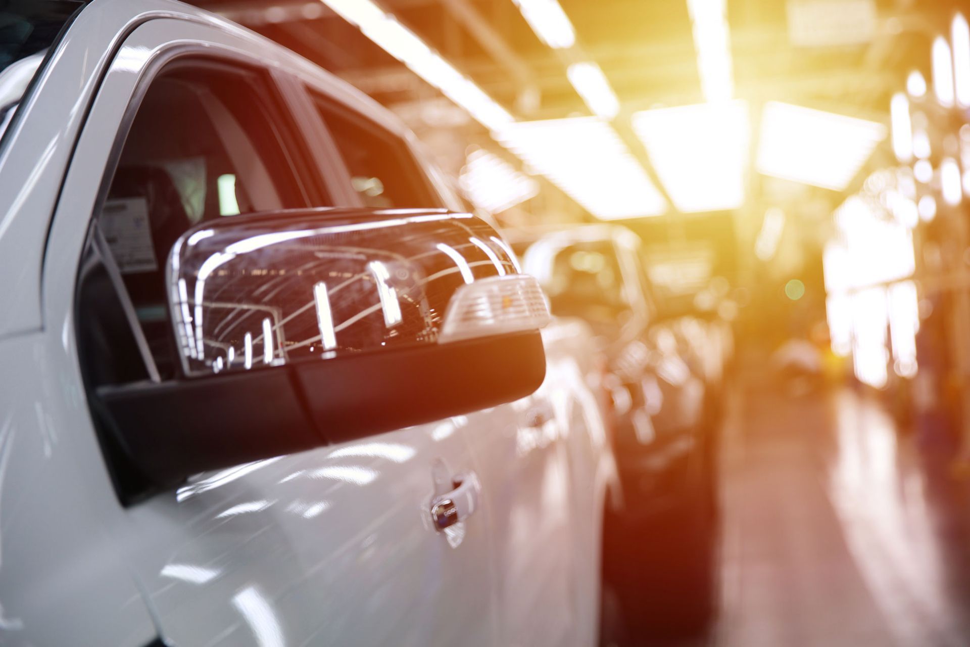 Ein weißes Auto auf einem Montageband, dessen Seitenspiegel von hellen Deckenleuchten reflektiert wird.