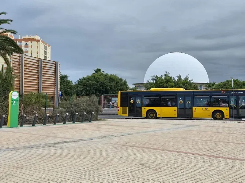 Autobús amarillo estacionado frente a un gran edificio de cúpula blanca y una estructura de madera, bajo un cielo nublado.