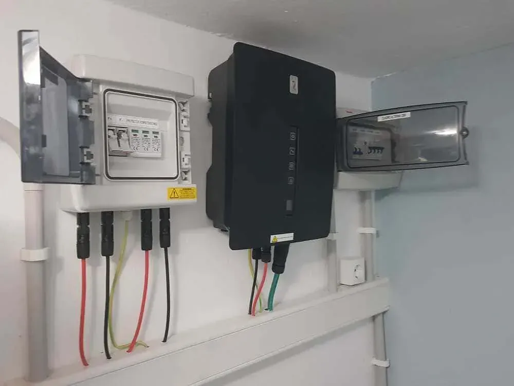 Equipo de panel solar montado en una pared blanca, que incluye una caja de fusibles, un inversor y componentes eléctricos con cables.