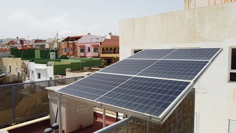 Paneles solares instalados en un tejado de una zona residencial soleada. Los paneles son negros y están orientados hacia el cielo.
