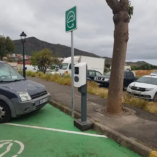 Estación de carga de vehículos eléctricos con plaza de aparcamiento verde y cartel, una furgoneta gris estacionada cerca.