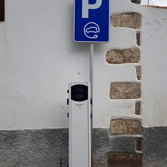 Estación de carga de vehículos eléctricos con una señal de estacionamiento azul frente a una pared blanca.