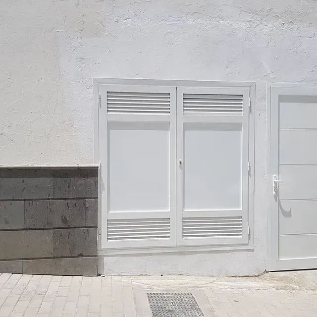 Ventana blanca con lamas horizontales en una pared blanca. A la izquierda y a la derecha, respectivamente, en la acera, hay un ladrillo gris y una puerta.