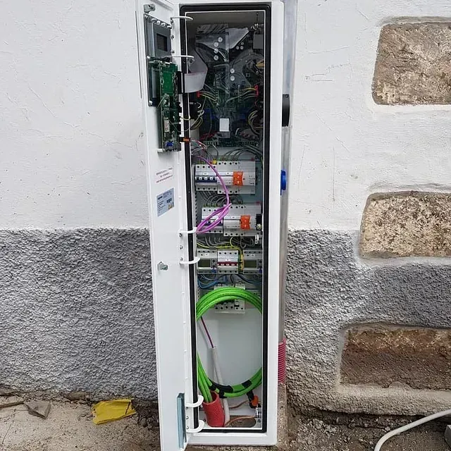 Armario eléctrico blanco abierto, montado en la pared, que muestra el cableado, los disyuntores y un cable verde. Ubicado en el exterior, junto a una pared de piedra texturizada.
