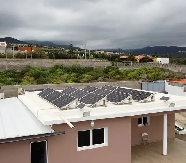 Paneles solares en el tejado de una casa de color claro contra un cielo nublado. Al fondo, vegetación exuberante y montañas lejanas.