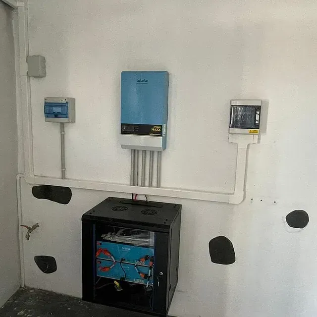 Una sala eléctrica con equipo de paneles solares montado en una pared blanca, que incluye un inversor azul y un bastidor de equipo negro.