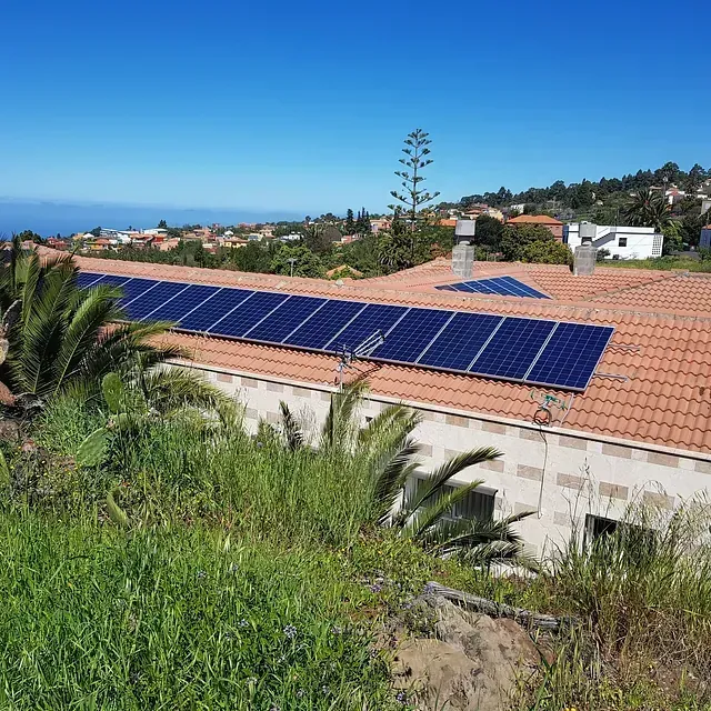 Paneles solares sobre un tejado de tejas rojas con vistas a un pueblo costero soleado con follaje verde.