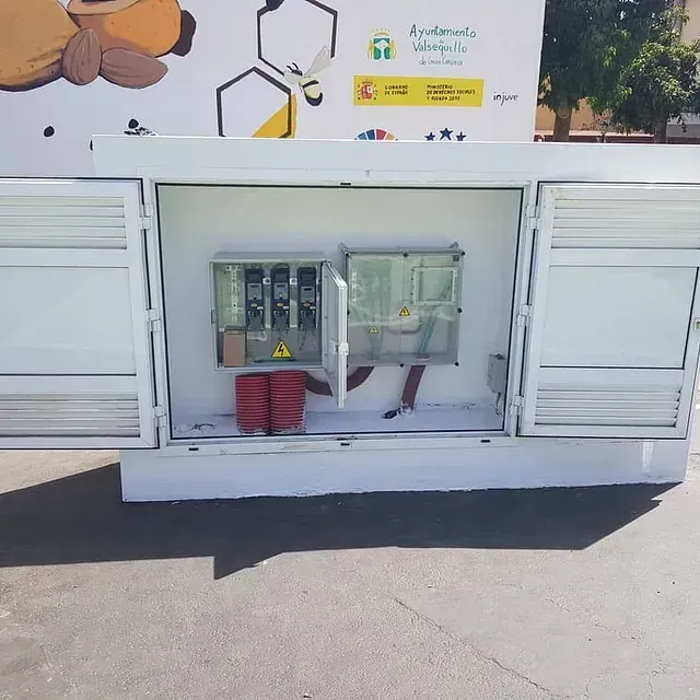 Armario eléctrico blanco abierto con las puertas entreabiertas sobre una superficie de hormigón. En su interior, hay equipos eléctricos y conos de seguridad rojos.