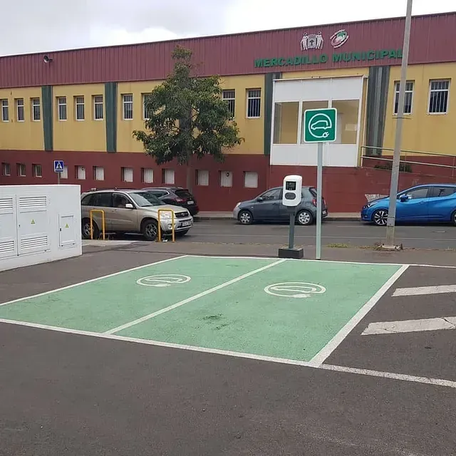 Estación de carga de vehículos eléctricos frente a un edificio municipal; puntos de carga verdes, vehículos estacionados cerca y una estación de carga.