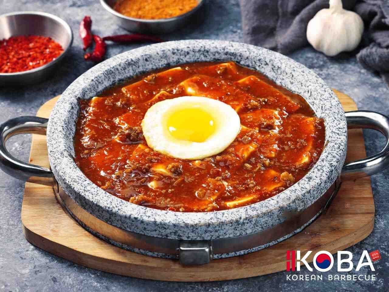 Guiso de tofu en recipiente de piedra cubierto con huevo frito, servido con hojuelas de chile, condimentos y ajo.