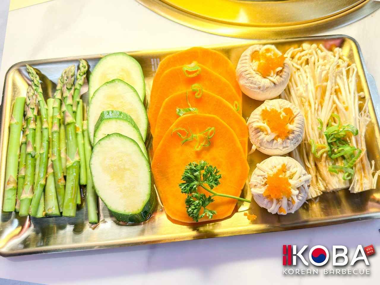 Plato de verduras: espárragos, calabacines, rodajas de calabaza, sombreros de champiñones y hongos enoki, en una bandeja dorada.