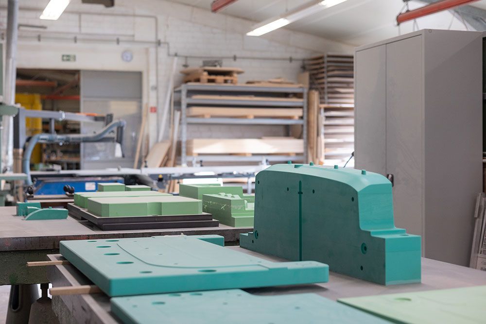 Grüne Schaumstoffformen auf einer Werkbank in einer Werkstatt. Im Hintergrund sind ein Schrank und Regale aus Holz zu sehen.