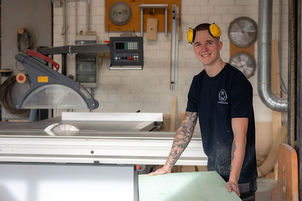 Ein Mann mit Gehörschutz lächelt in einer Werkstatt und hält Material in der Nähe einer Säge.