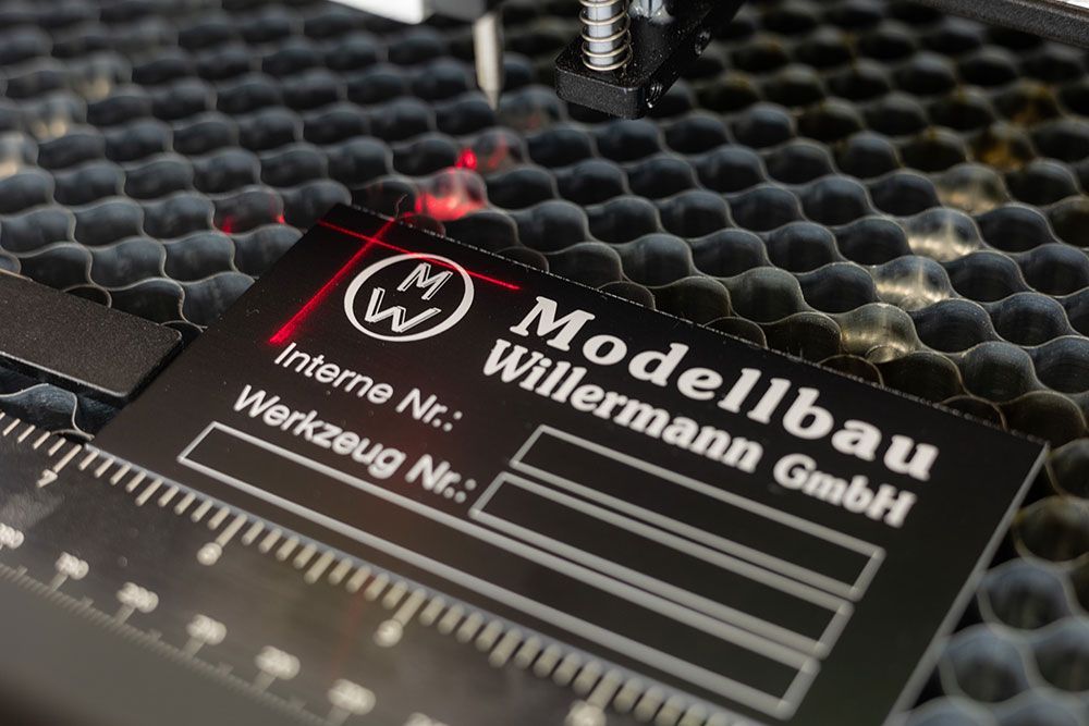 Lasergravur eines schwarzen Modelbau Willermann GmbH-Etiketts mit rotem Laser und wabenförmigem Hintergrund.