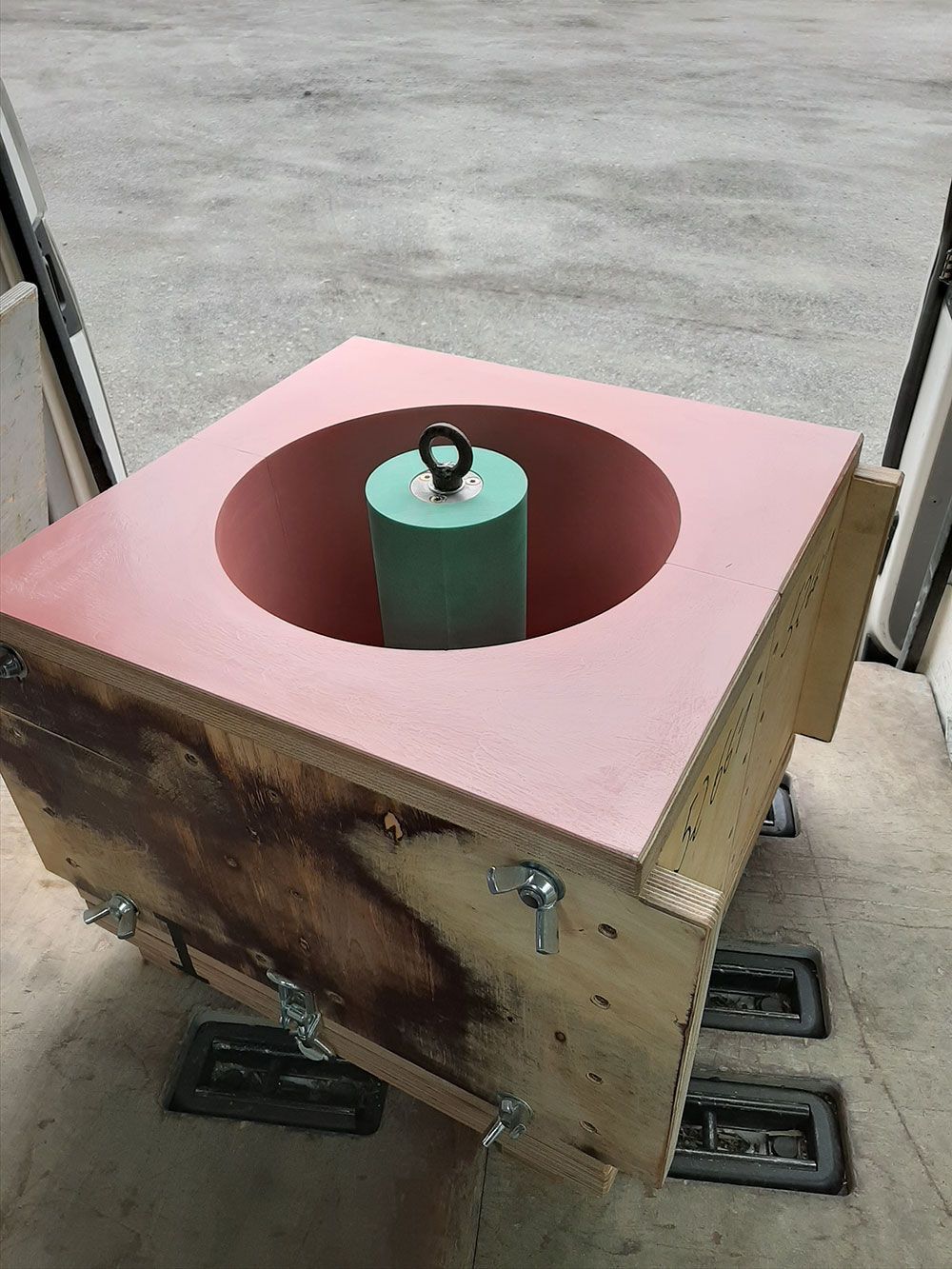 Holzkiste mit rosa umrandeter Öffnung, grünem Zylinder im Inneren, Details aus Metallverschluss.