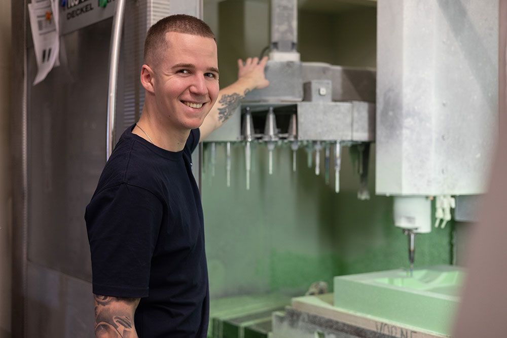 Ein Mann lächelt und zeigt auf eine CNC-Maschine in einer Werkstatt. Die Maschine bohrt in einen grünen Block.
