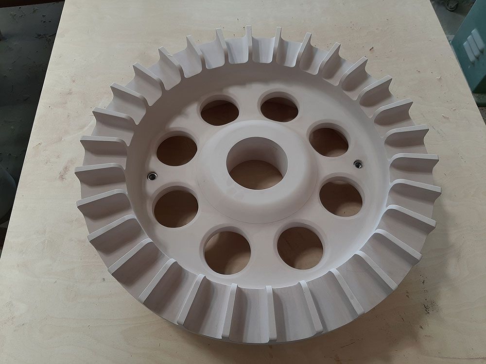 Großes, kreisrundes, cremefarbenes mechanisches Bauteil mit radialen Rippen und zentralen sowie peripheren Löchern.