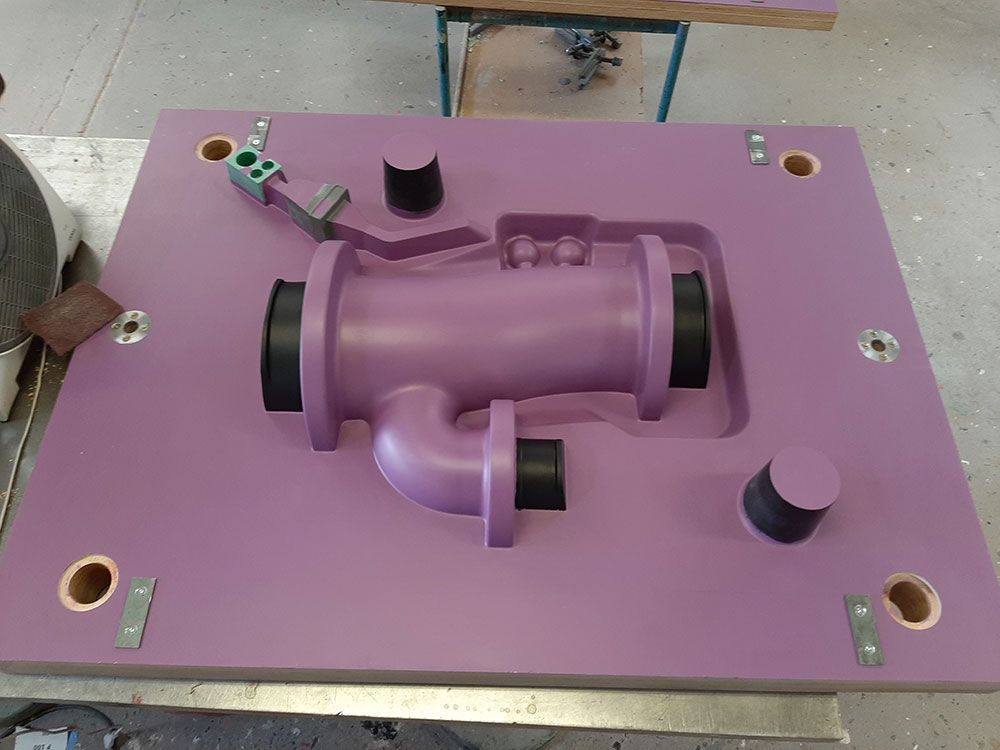 Lila Gussform mit einem Rohrelement, schwarzen Stopfen und verschiedenen Metallstützen auf einer Arbeitsfläche.