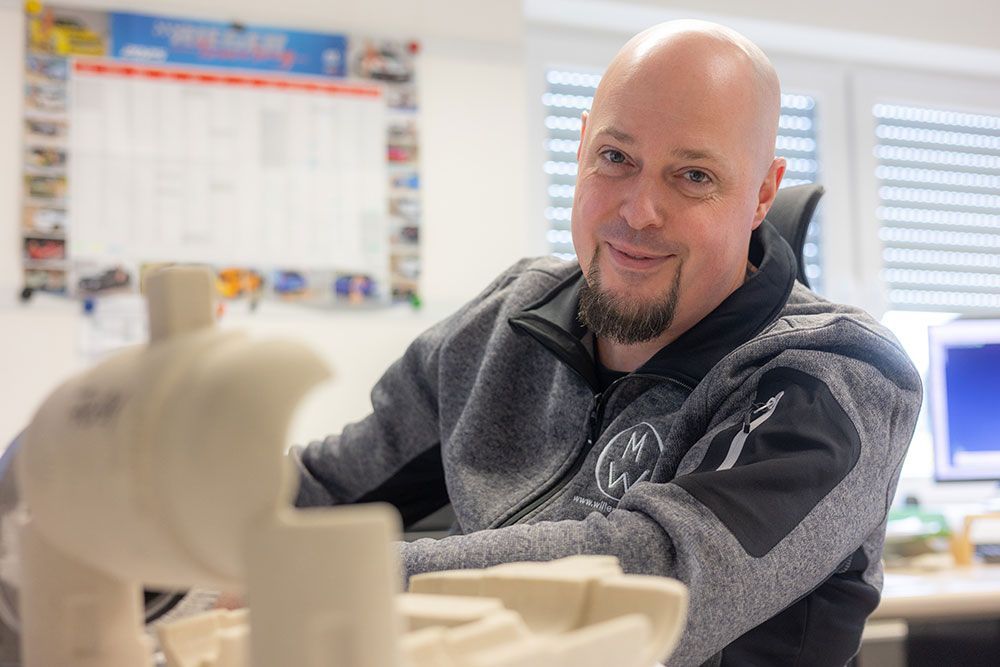 Glatzköpfiger Mann mit Spitzbart lächelt, sitzt mit einem Model an einem Schreibtisch, Bürohintergrund.
