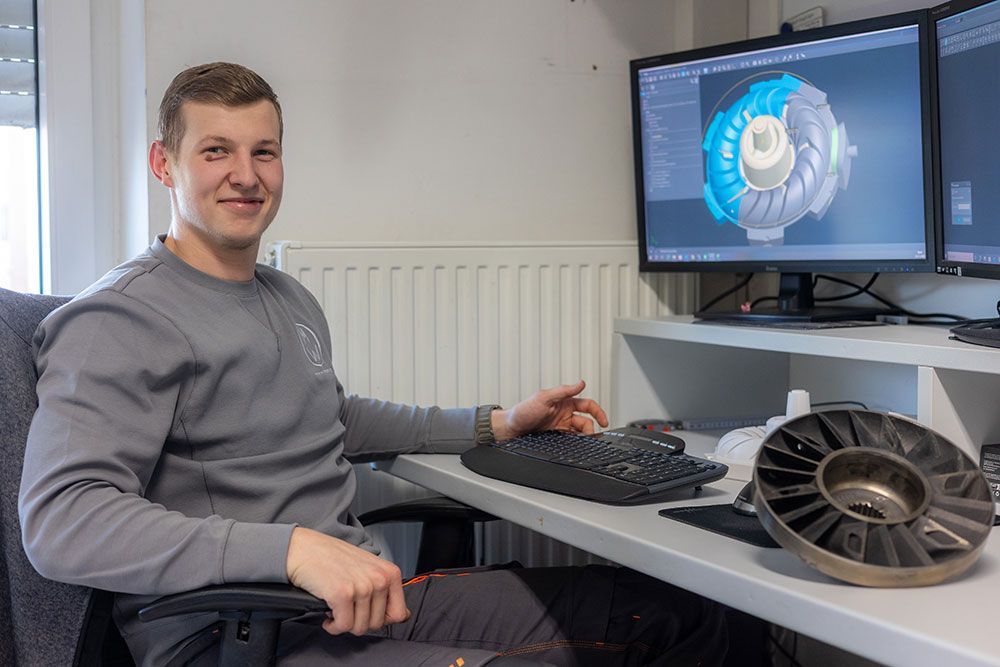 Mann sitzt lächelnd an einem Schreibtisch mit Computer und einem Bauteil. Auf dem Bildschirm wird die Konstruktion gezeigt.