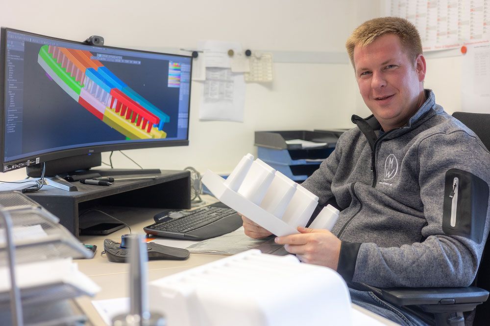 Ein Mann hält ein weißes, 3D-gedrucktes Teil in der Hand und lächelt in die Kamera. Er befindet sich in einem Büro.