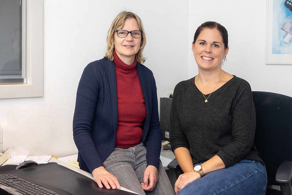 Zwei lächelnde Frauen sitzen an einem Schreibtisch in einem Büro.