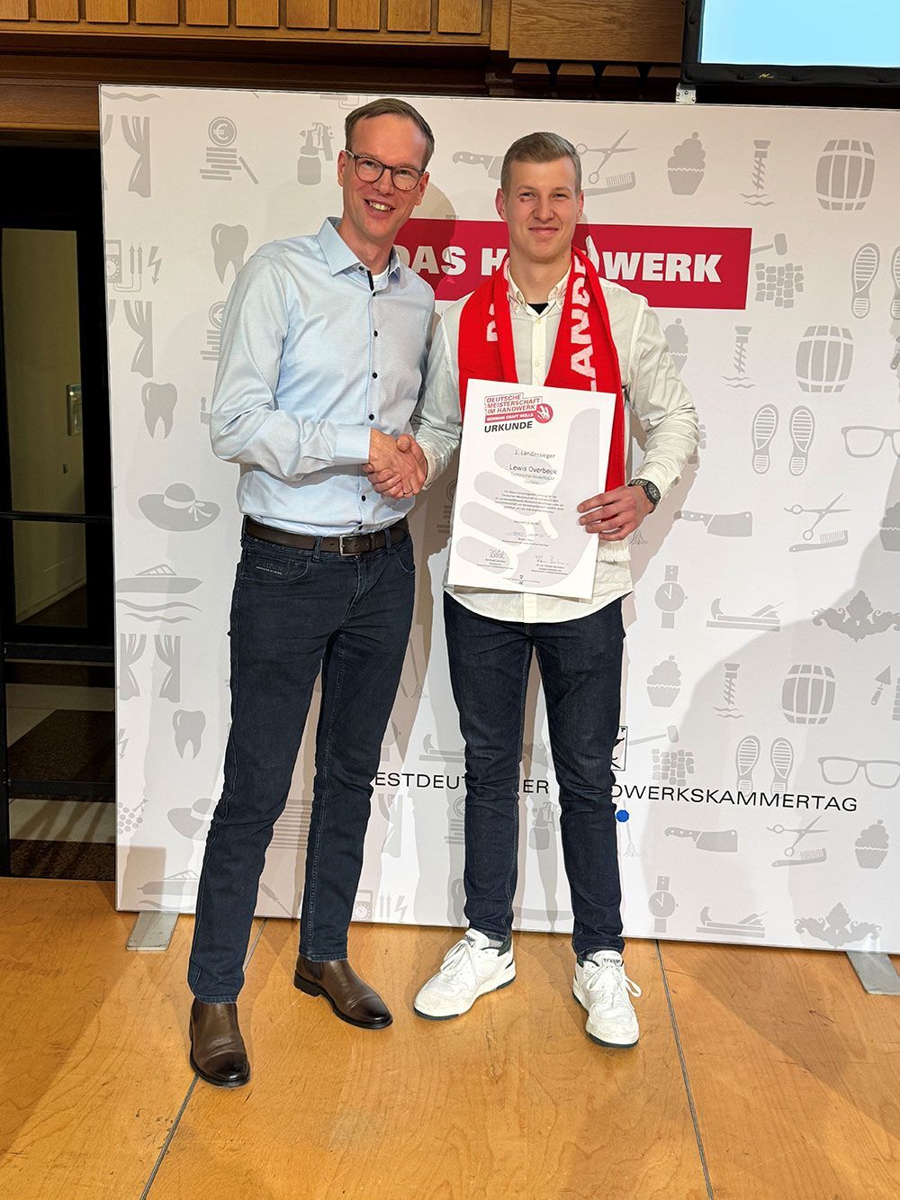 Zwei Männer stehen nebeneinander, einer schüttelt dem anderen die Hand. Der Mann rechts hält eine Urkunde in der Hand.