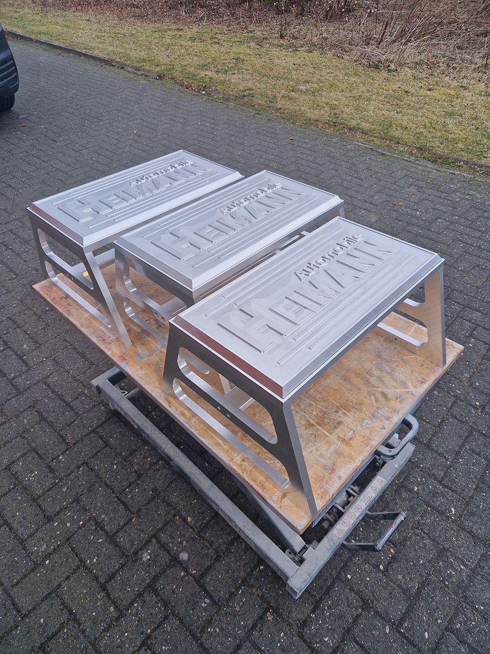 Drei metallene Tritthocker auf einer Holzplattform über einem grauen Metalllift, im Freien auf dem Gehweg.