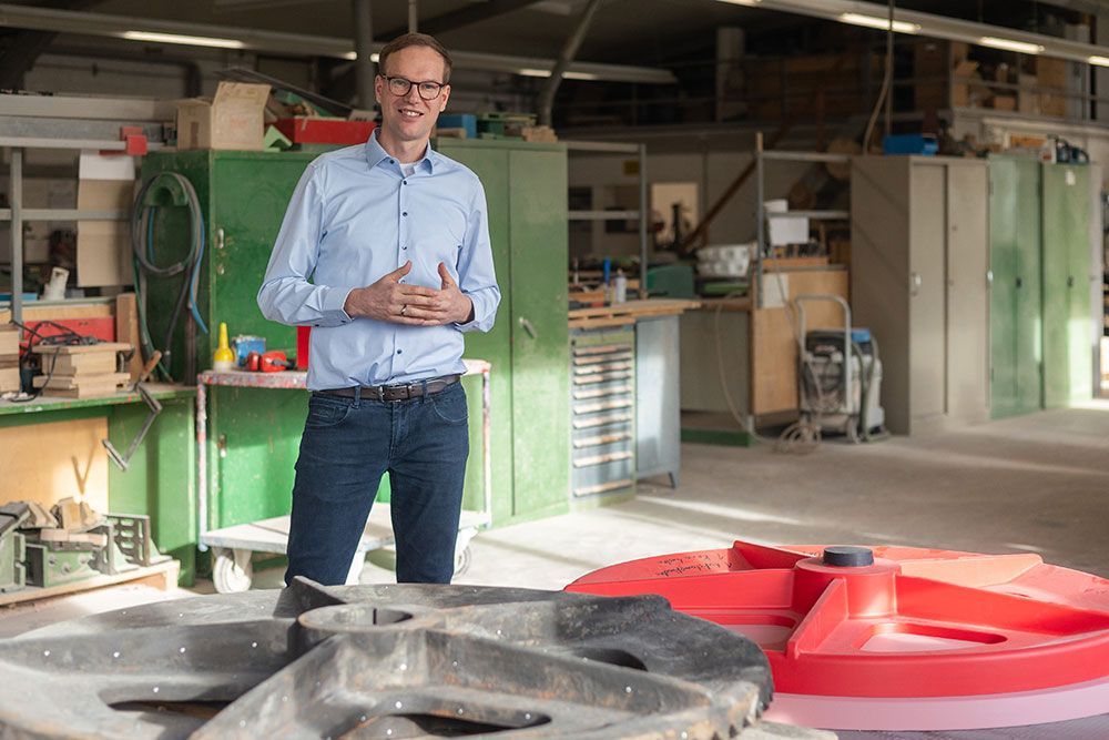 Ein Mann in blauem Hemd und Jeans lächelt in einer Werkstatt; im Vordergrund sind verschiedene Objekte zu sehen.