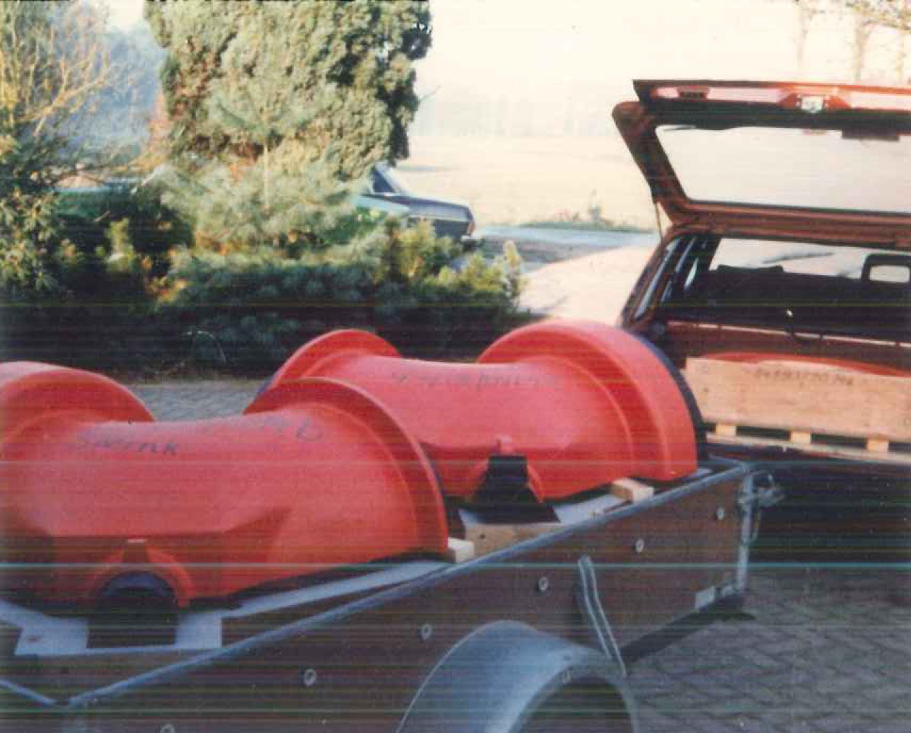 Zwei große rote Rohre auf einem Anhänger, daneben ein offener Kofferraum, im Freien.