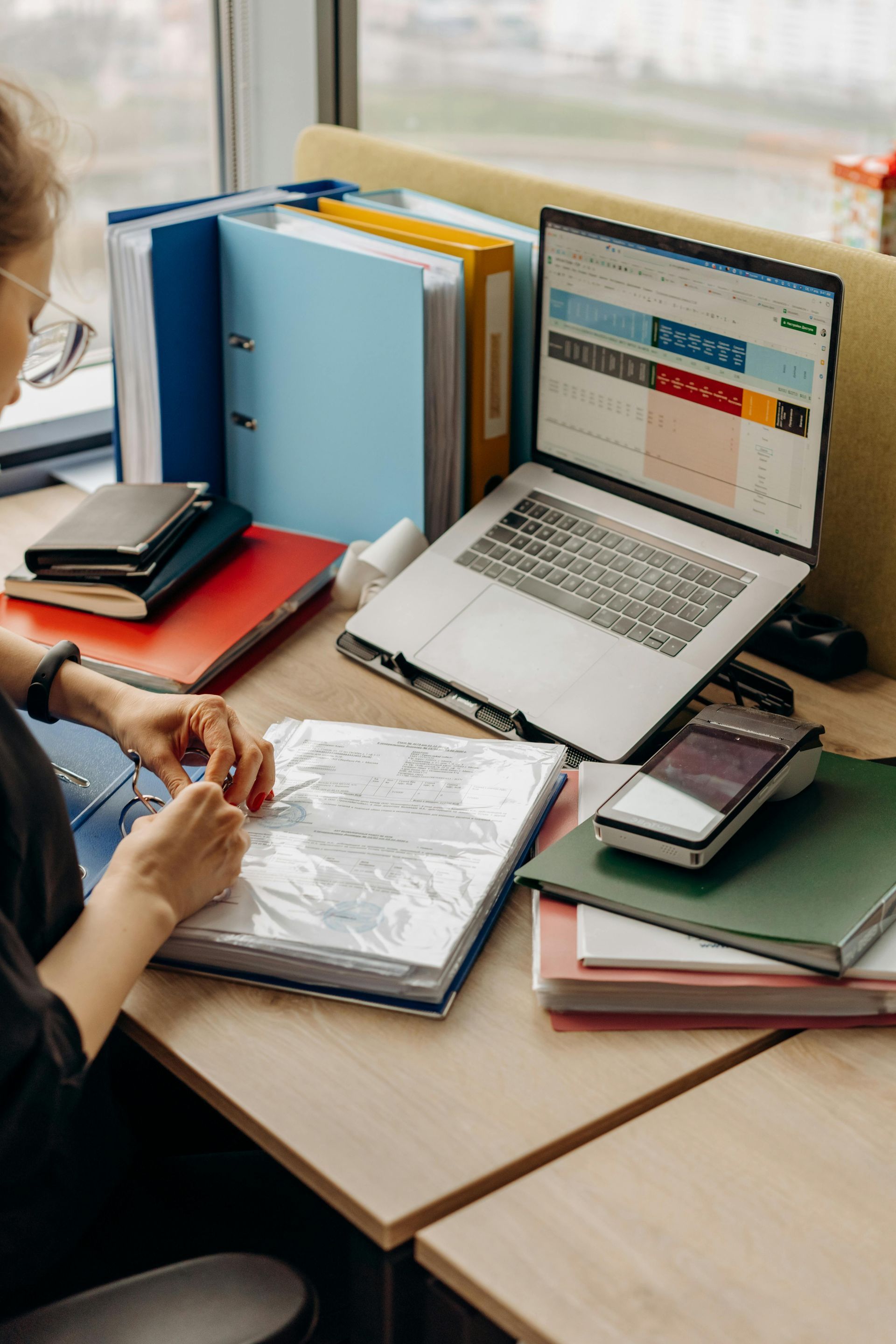 Une personne travaille à un bureau avec un ordinateur portable, des dossiers, des carnets et des papiers.