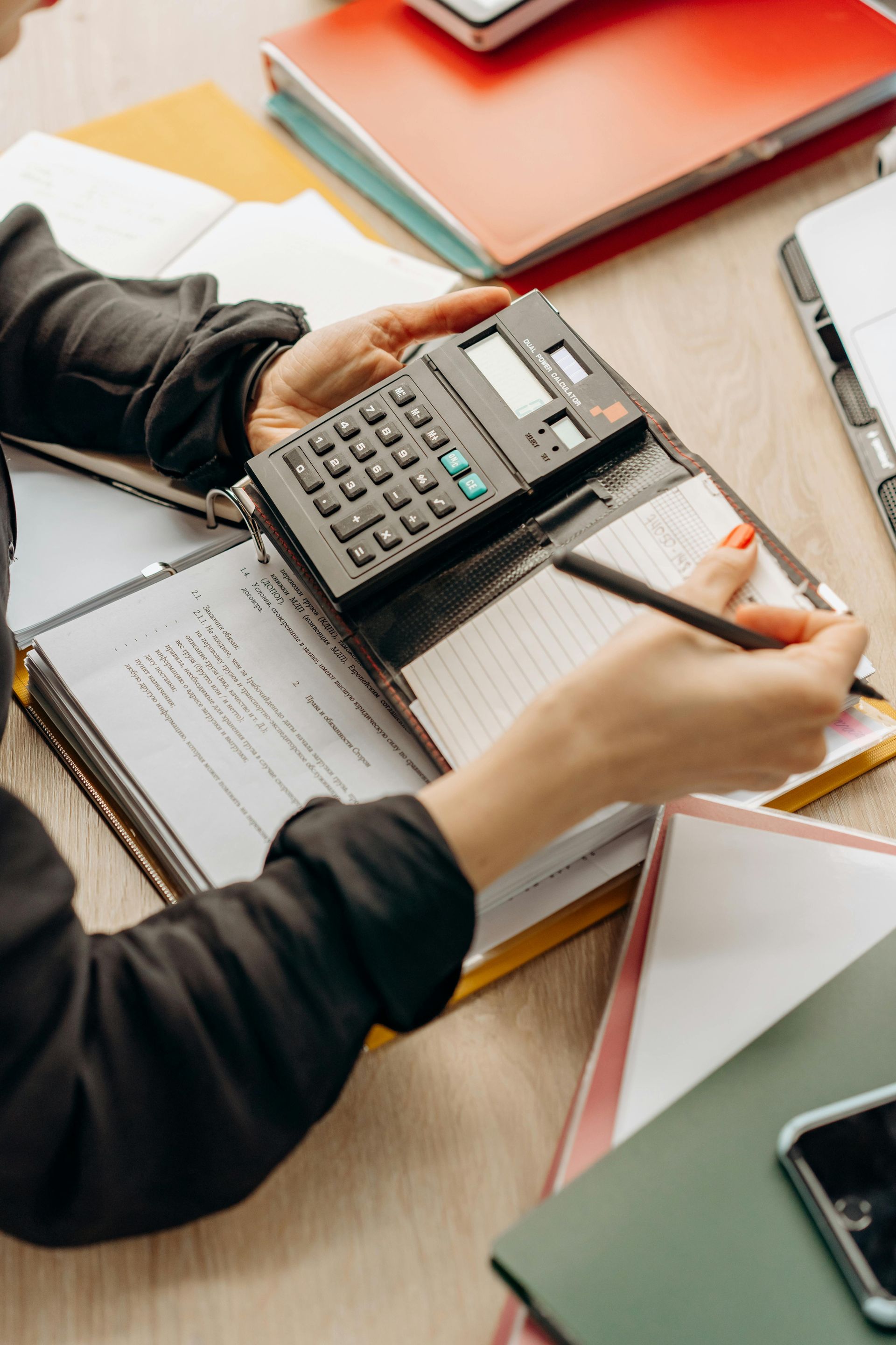 Des mains manipulant une calculatrice et un stylo au-dessus de documents posés sur un bureau recouvert de dossiers.