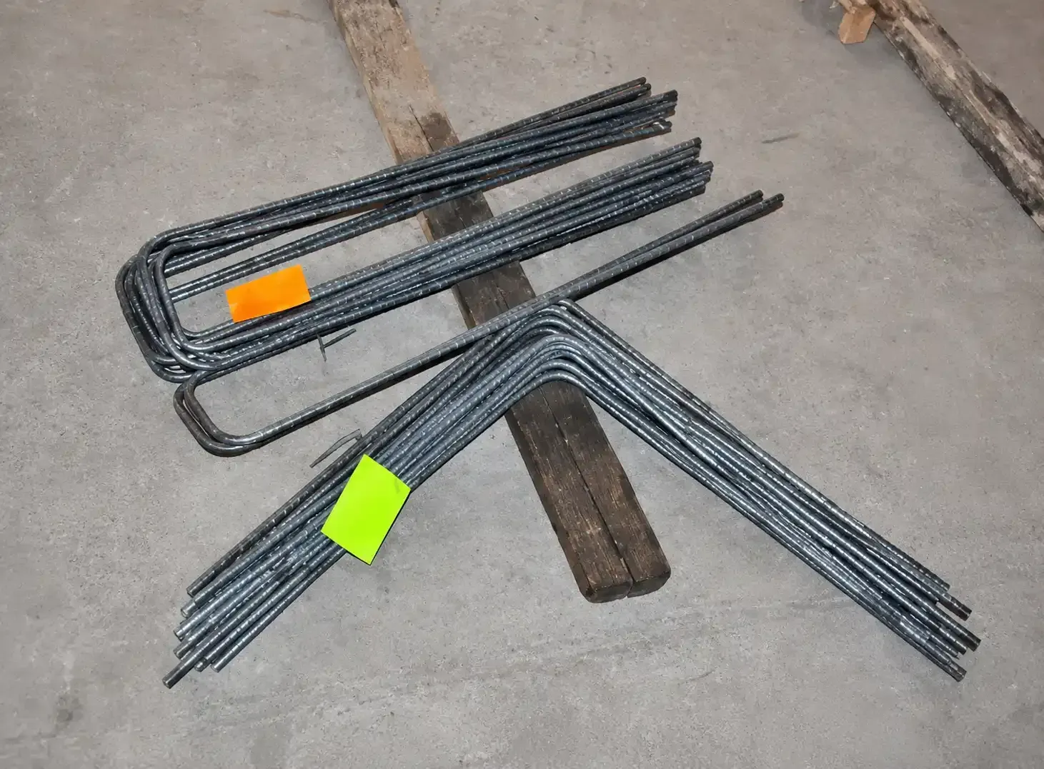 Barras de metal negro apiladas con etiquetas naranjas y verdes sobre un suelo de hormigón.