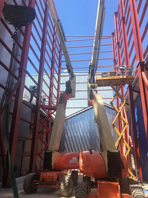 Dos plataformas elevadoras naranjas dentro de un edificio con estructura de acero en construcción. Se ven vigas rojas y cielo azul.