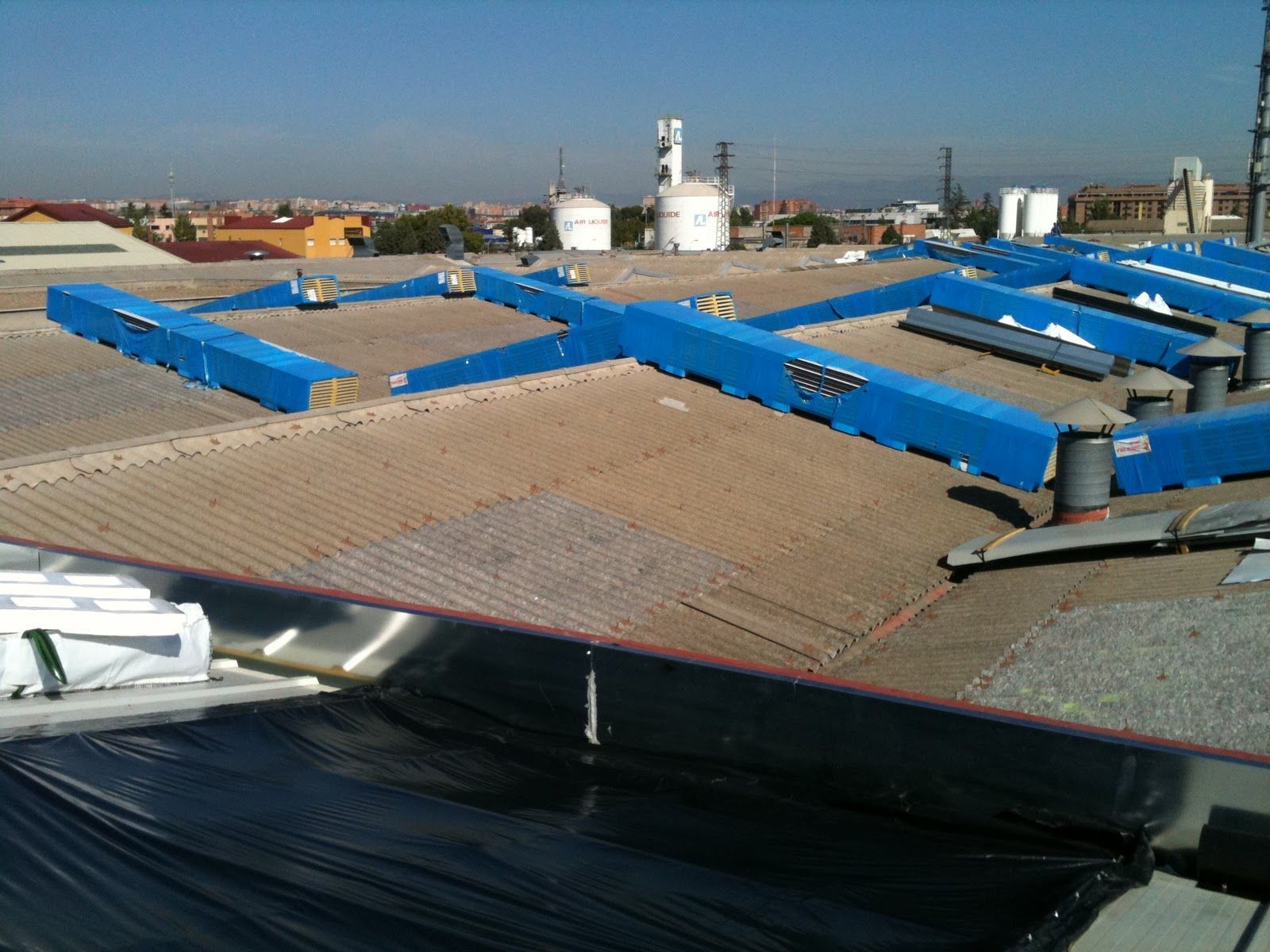 Conductos de HVAC azules sobre un techo corrugado de un edificio con otras estructuras al fondo.