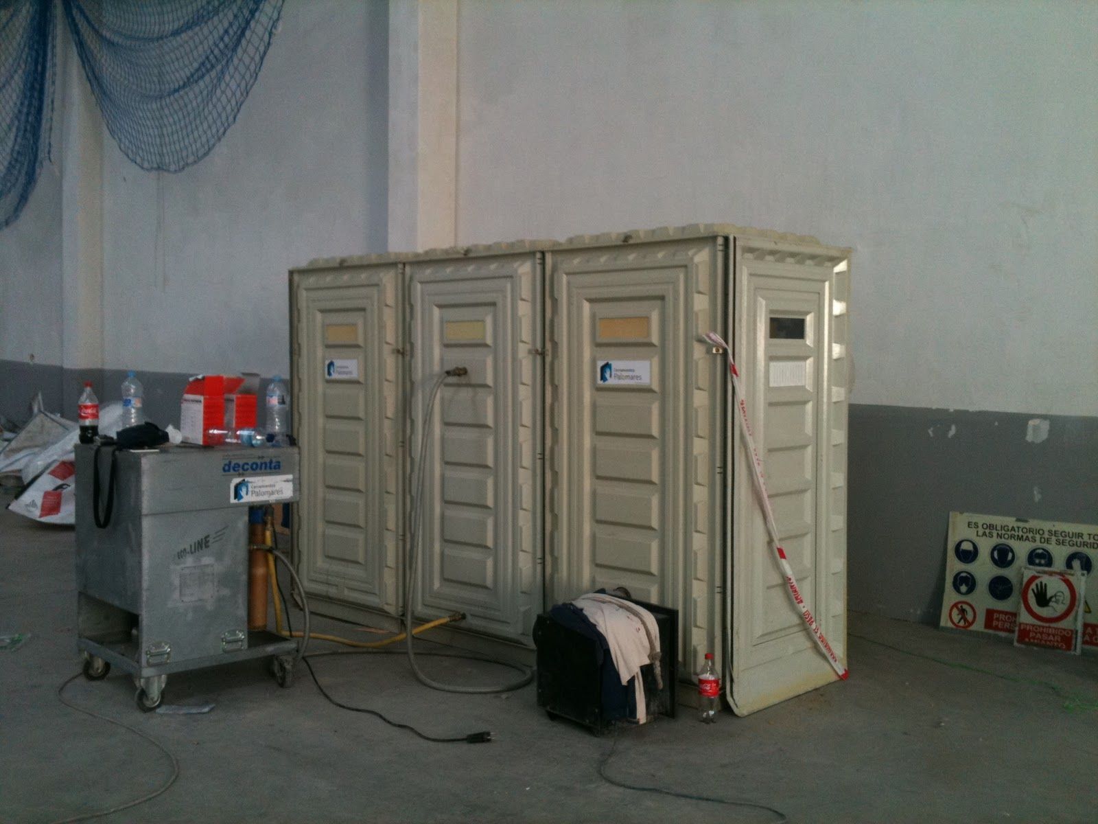 Tres baños portátiles dentro de un edificio con un carrito de metal y suministros cerca.