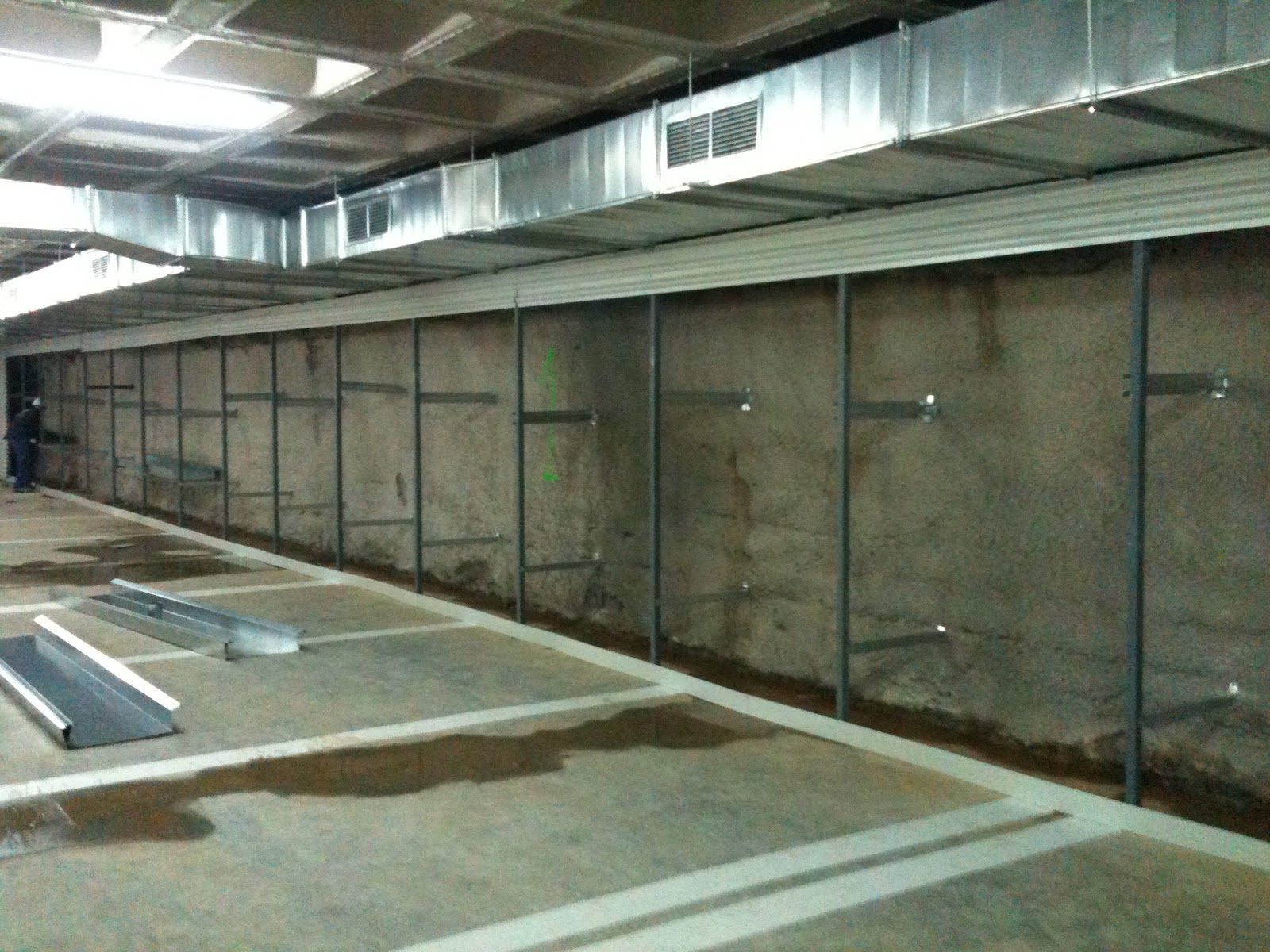 Habitación de hormigón con estructura metálica y conductos. Suelo húmedo con contornos rectangulares, posiblemente un aparcamiento.