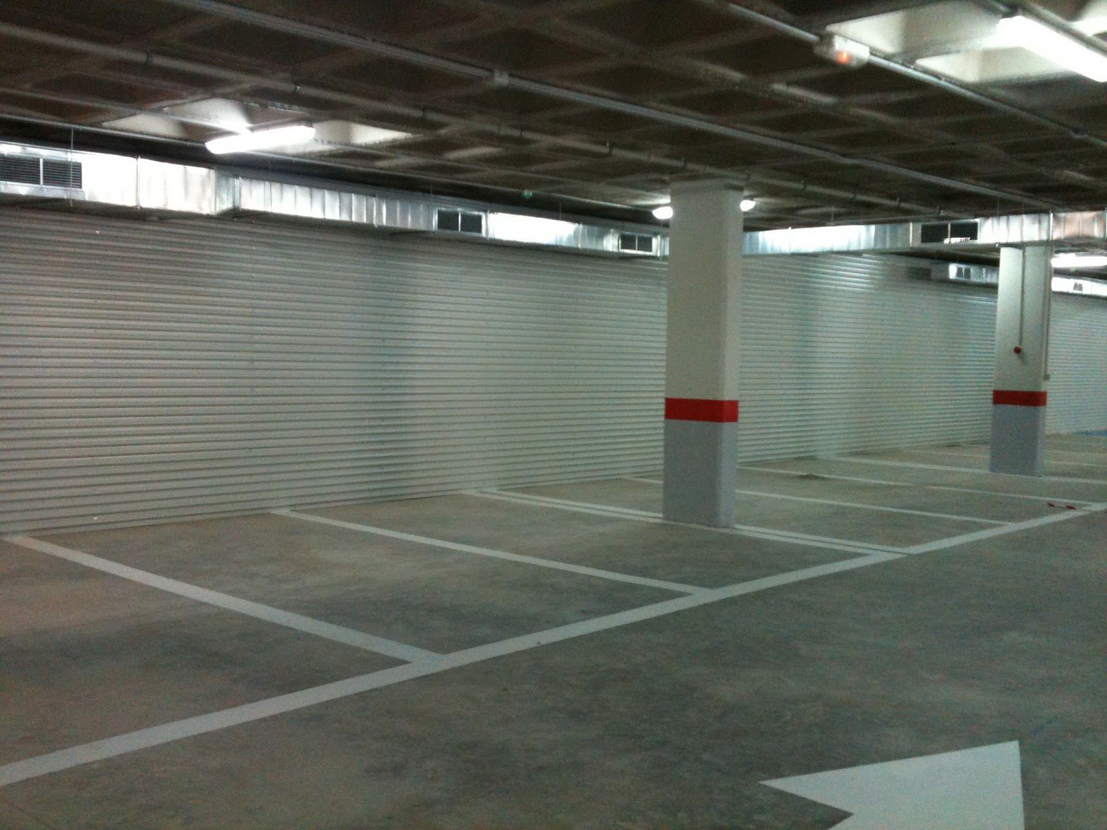 Plazas de aparcamiento vacías con líneas blancas, suelo de hormigón, pared metálica nervada y columnas de soporte con bandas rojas.