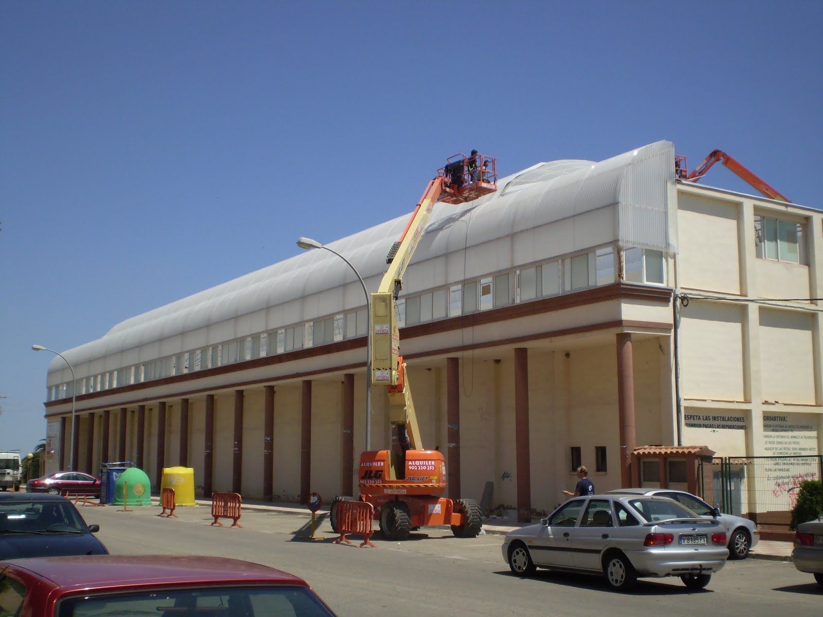 Edificio con tejado abovedado; un trabajador en un ascensor naranja repara el tejado bajo la luz del sol. Calle con coches en primer plano.