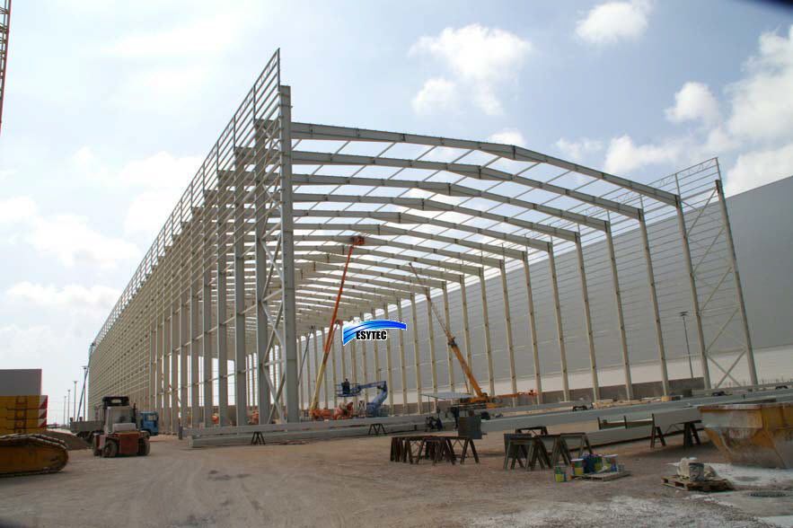 Obra con estructura de acero de un gran almacén. Grúas y maquinaria pesada bajo un cielo parcialmente nublado.
