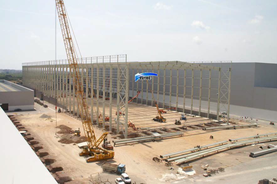 Sitio de construcción: Estructura de acero de un gran edificio en construcción, grúa en funcionamiento, equipos y materiales visibles.