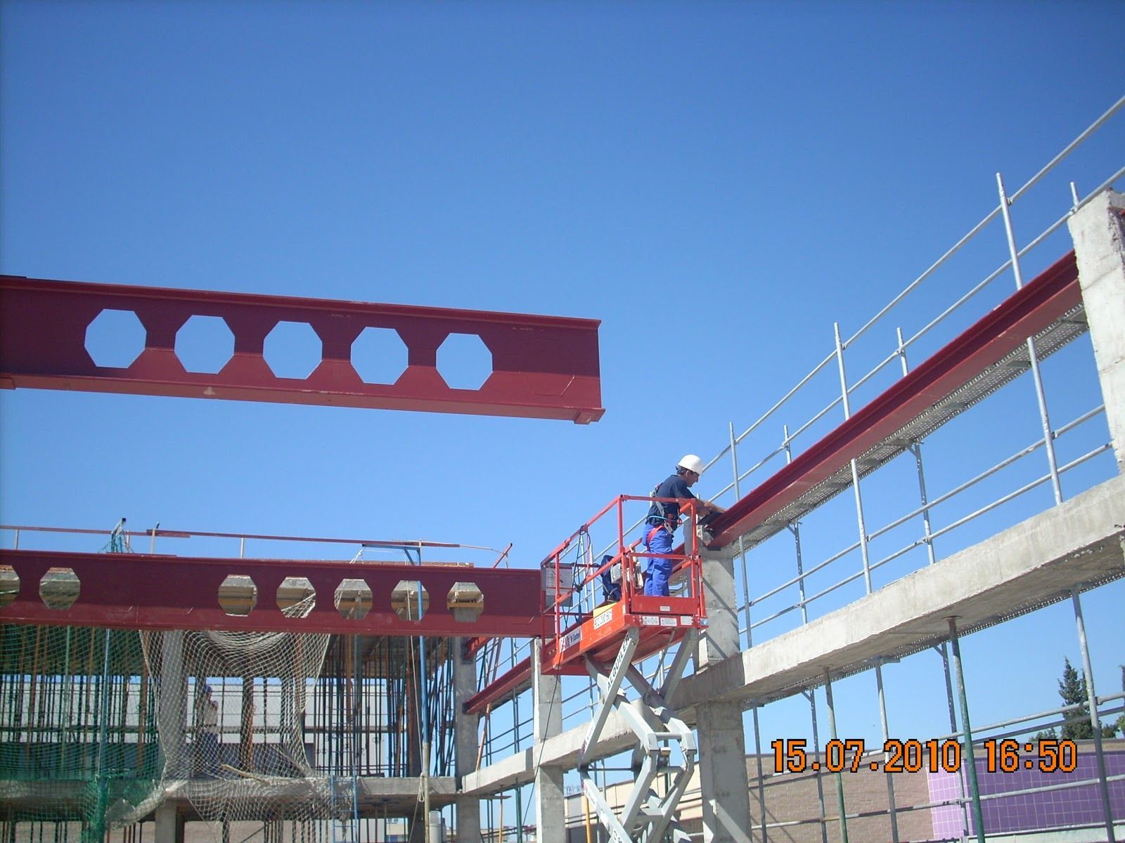 Trabajador de la construcción en un ascensor, conectando una viga de acero roja al edificio bajo un cielo azul.