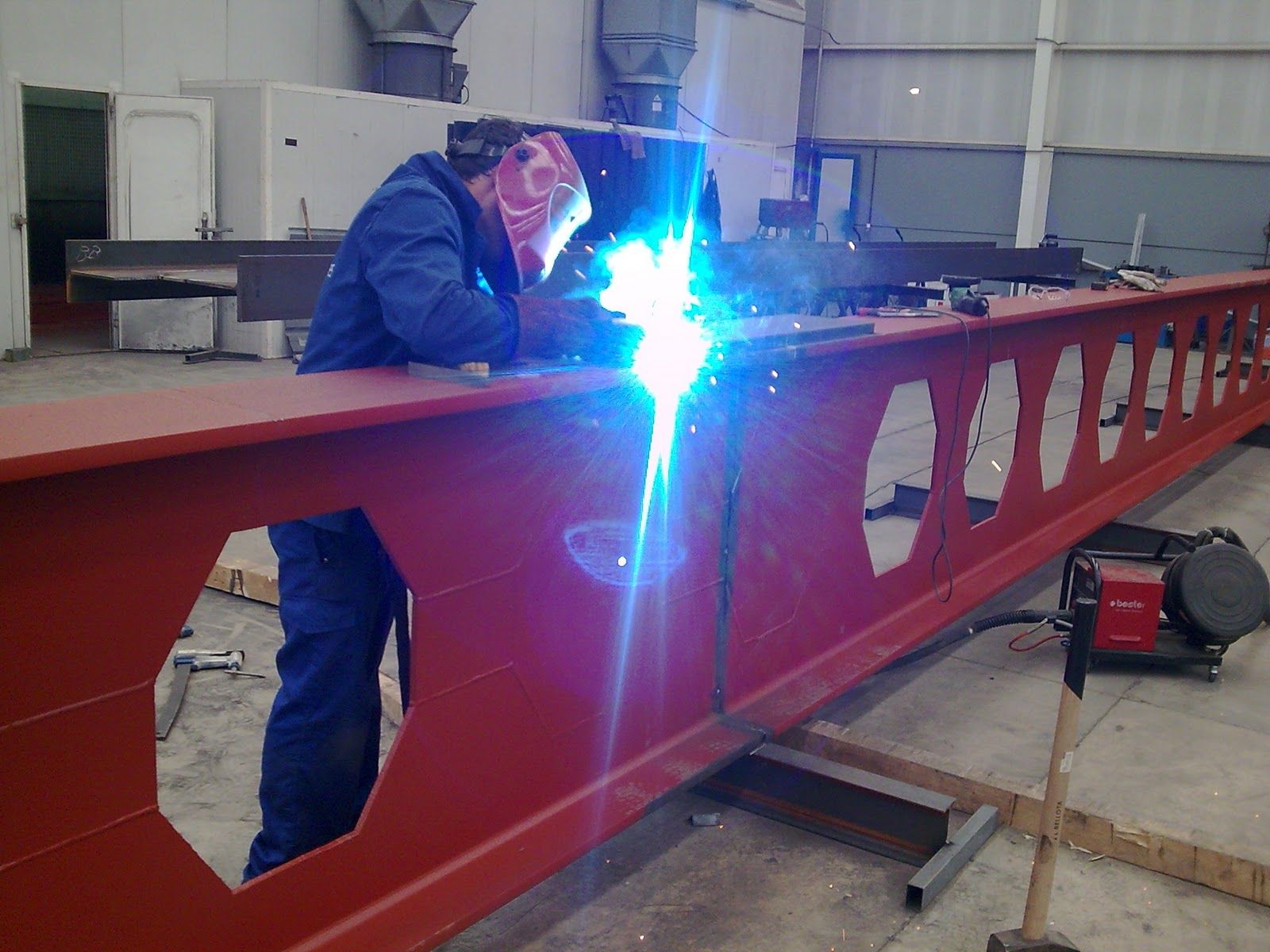 Soldador con mono azul usando un soplete sobre una viga metálica roja en el interior.