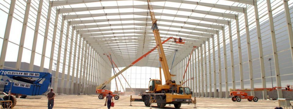 Obra de construcción con grúa amarilla y máquinas elevadoras dentro de un gran edificio con soportes metálicos.