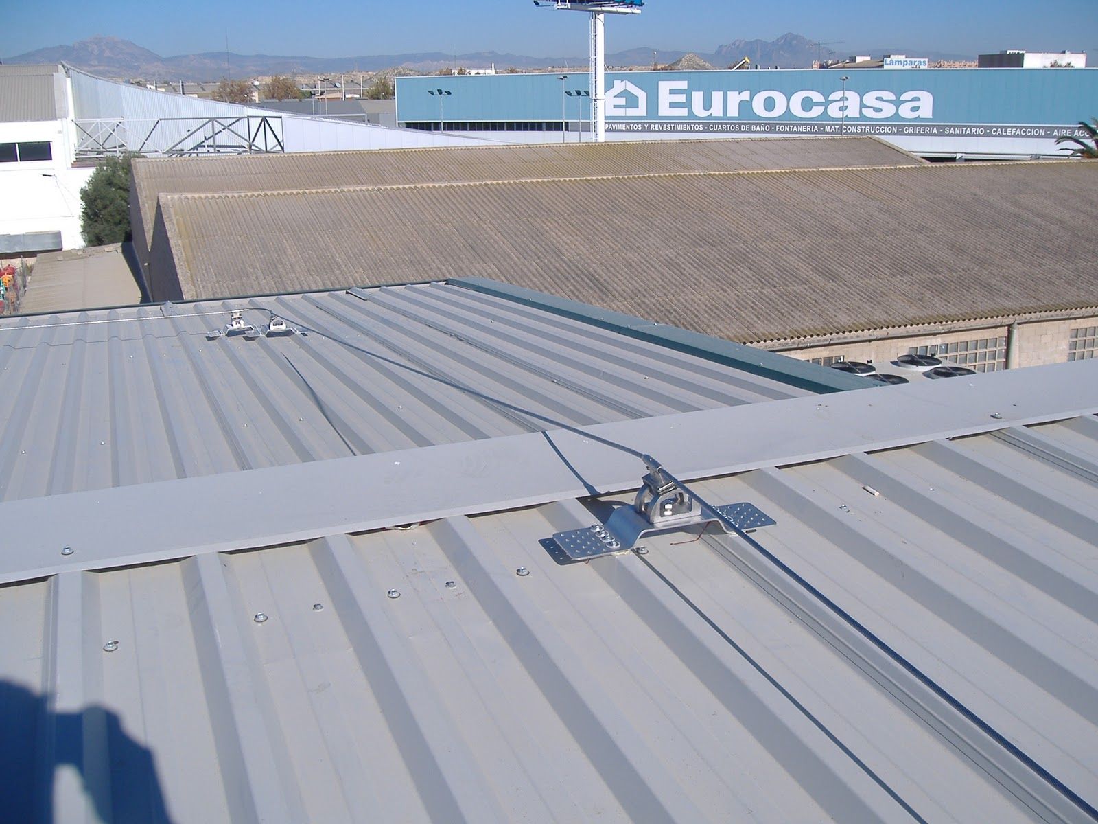 Techo de metal con cables de seguridad y anclajes, edificios industriales al fondo, día soleado.