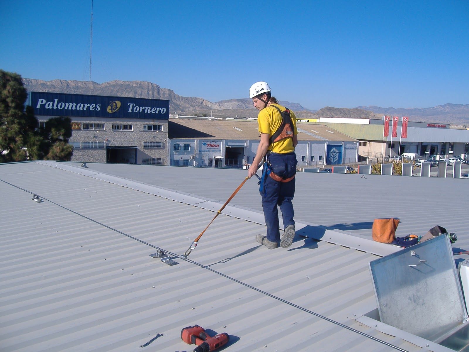 Trabajador en un tejado, asegurado con un arnés, inspeccionando un cable de seguridad. Día soleado, entorno industrial.